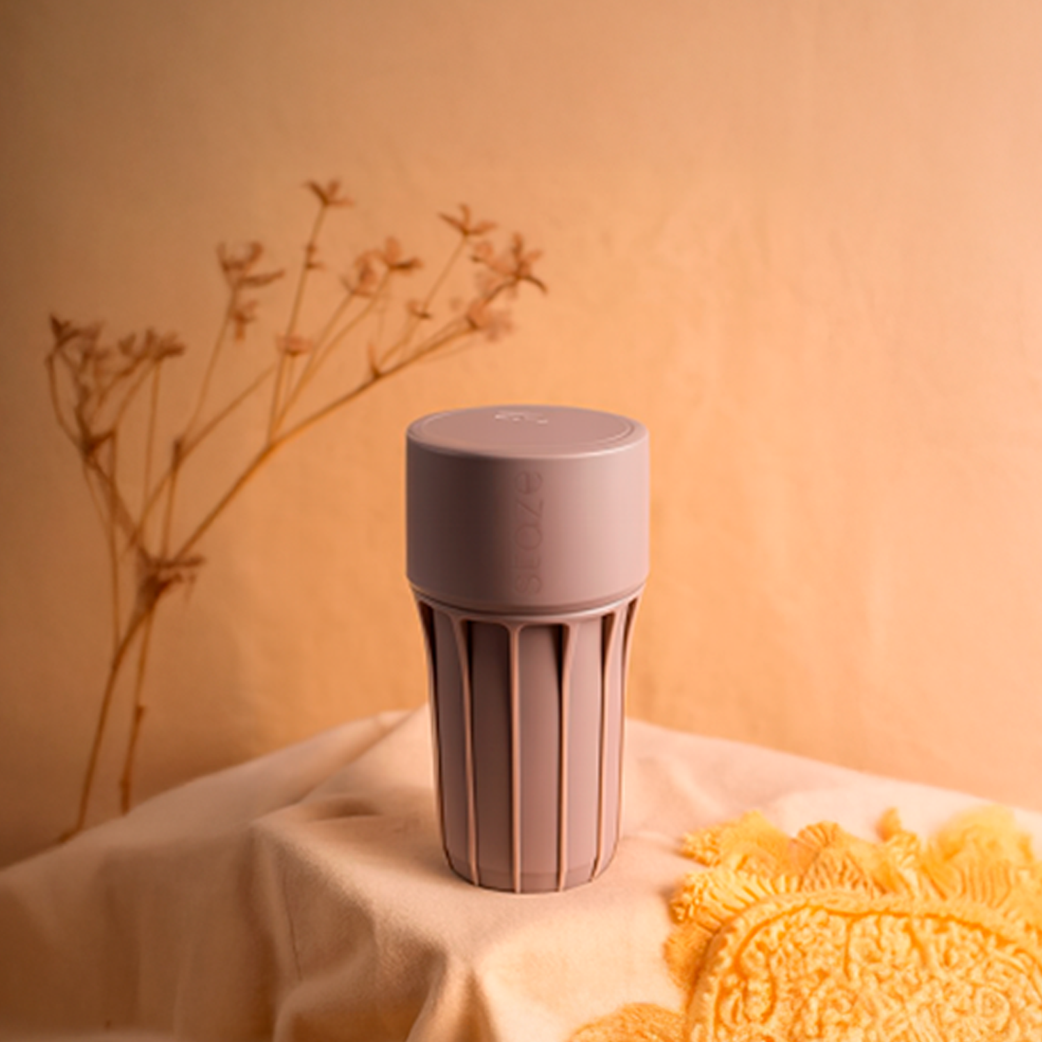 Soft-lit close-up of the Shroom-colored STAZE PRESERVE showing its cylindrical top and vertical ribbed body centered on a draped fabric with dried flowers blurred in the background