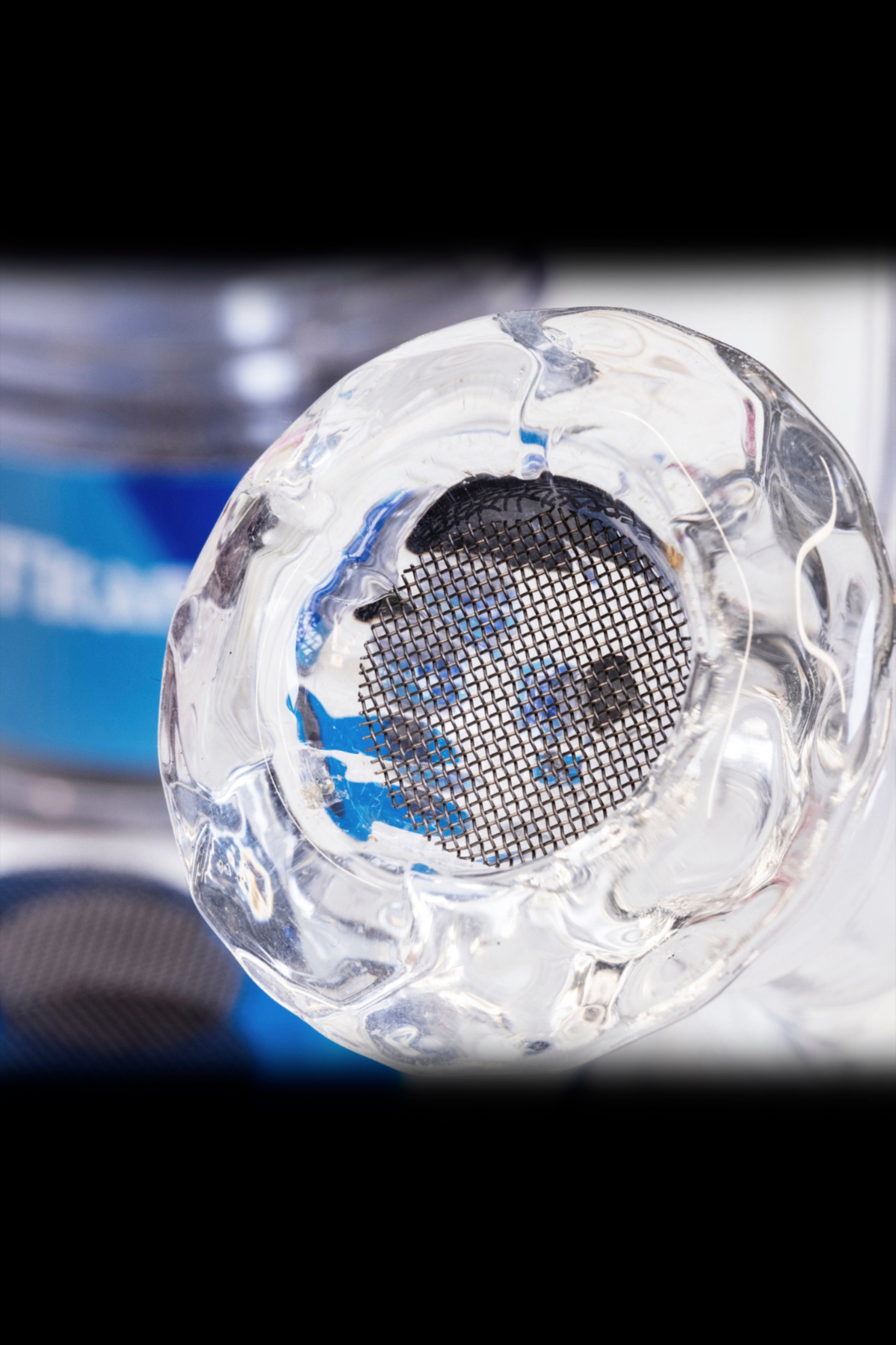 Clear glass bubbler with a stainless steel screen, blurred cannabis packaging in the background.