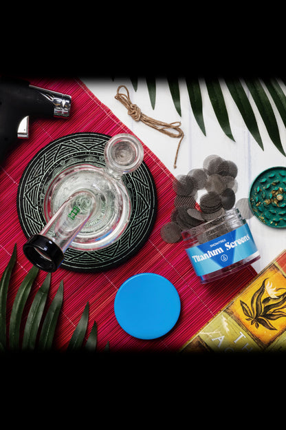 Overhead shot of cannabis accessories including a glass pipe, titanium screens, lighter, and rolling papers arranged on a patterned mat with tropical leaves.
