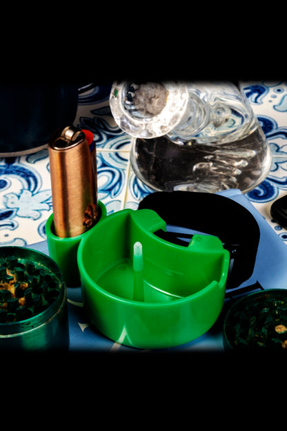 Cannabis accessories including a green grinder, small containers of bud, a lighter, and water bottle arranged on a patterned surface with dramatic lighting.