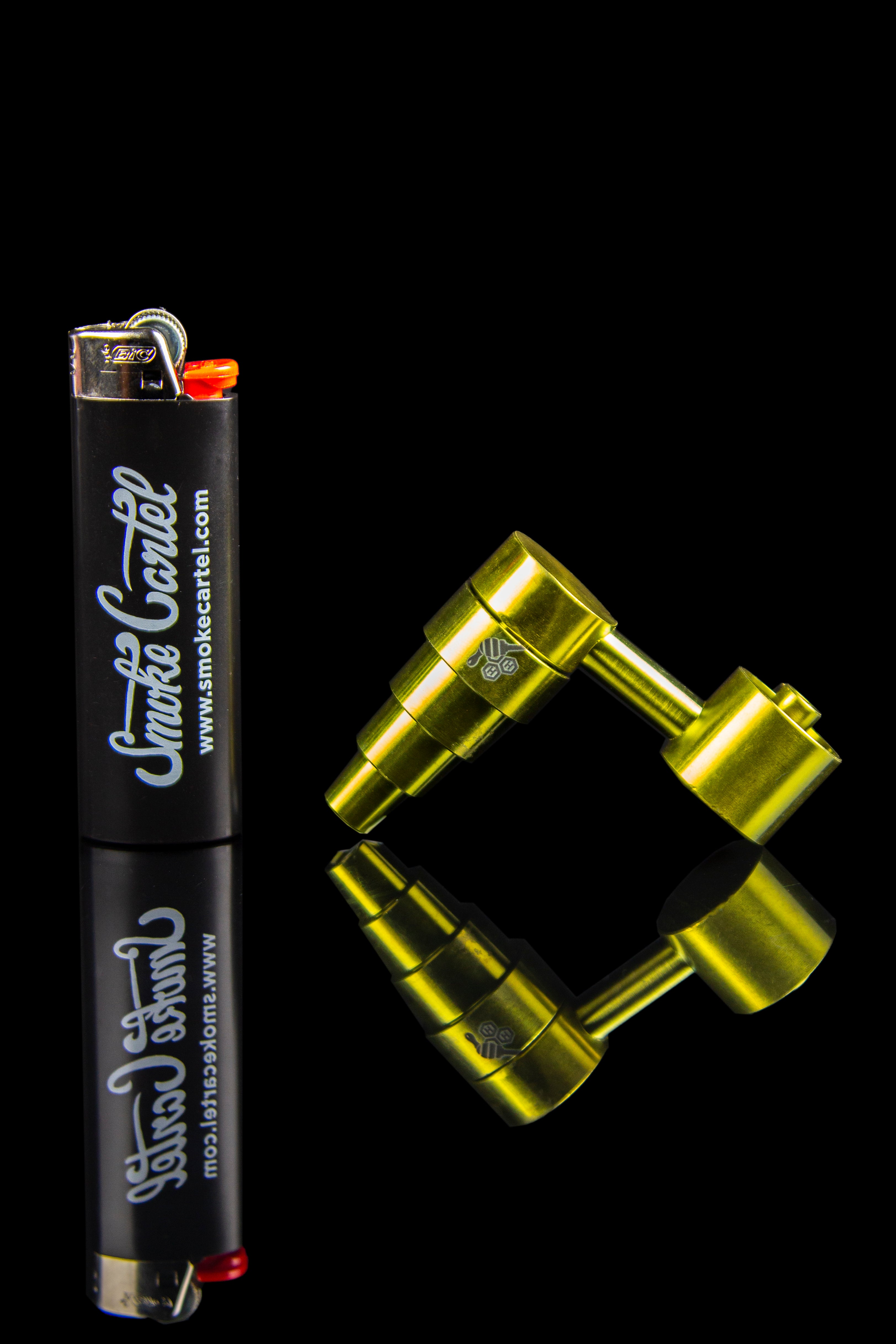 A black lighter with “Smoke Cartel” branding lies next to a gold cannabis oil rig on a black background.