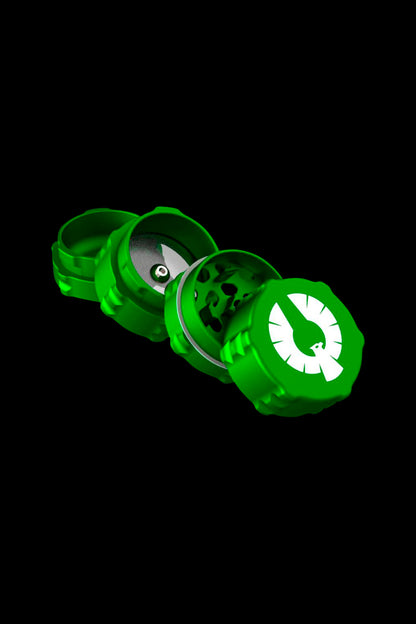 Green grinder with open chambers displaying ground cannabis, isolated on a black background.
