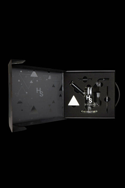 Open black gift box displaying cannabis smoking accessories, including a glass pipe, vaporizer, and tools, with a geometric pattern and "Hs" logo.