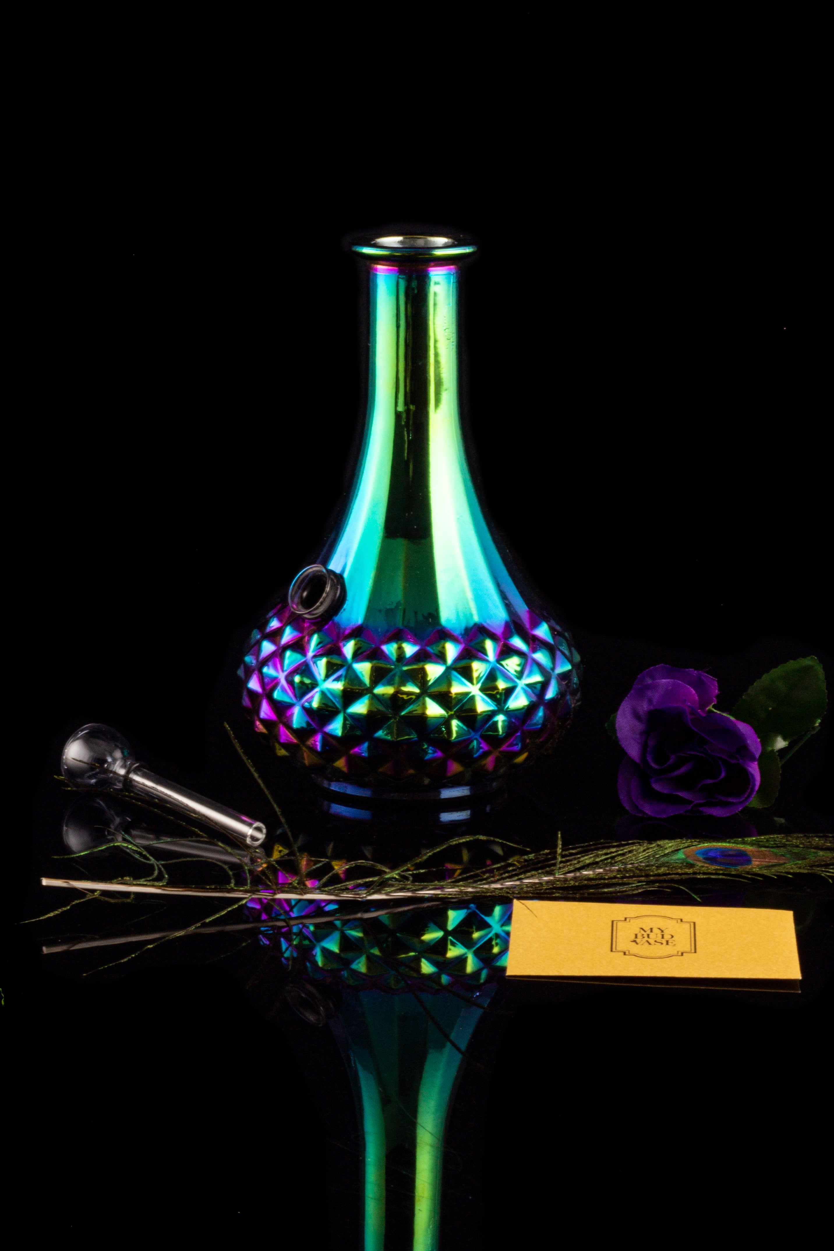 Iridescent glass water pipe with accompanying smoking tools and a purple rose, reflected on a mirrored surface.