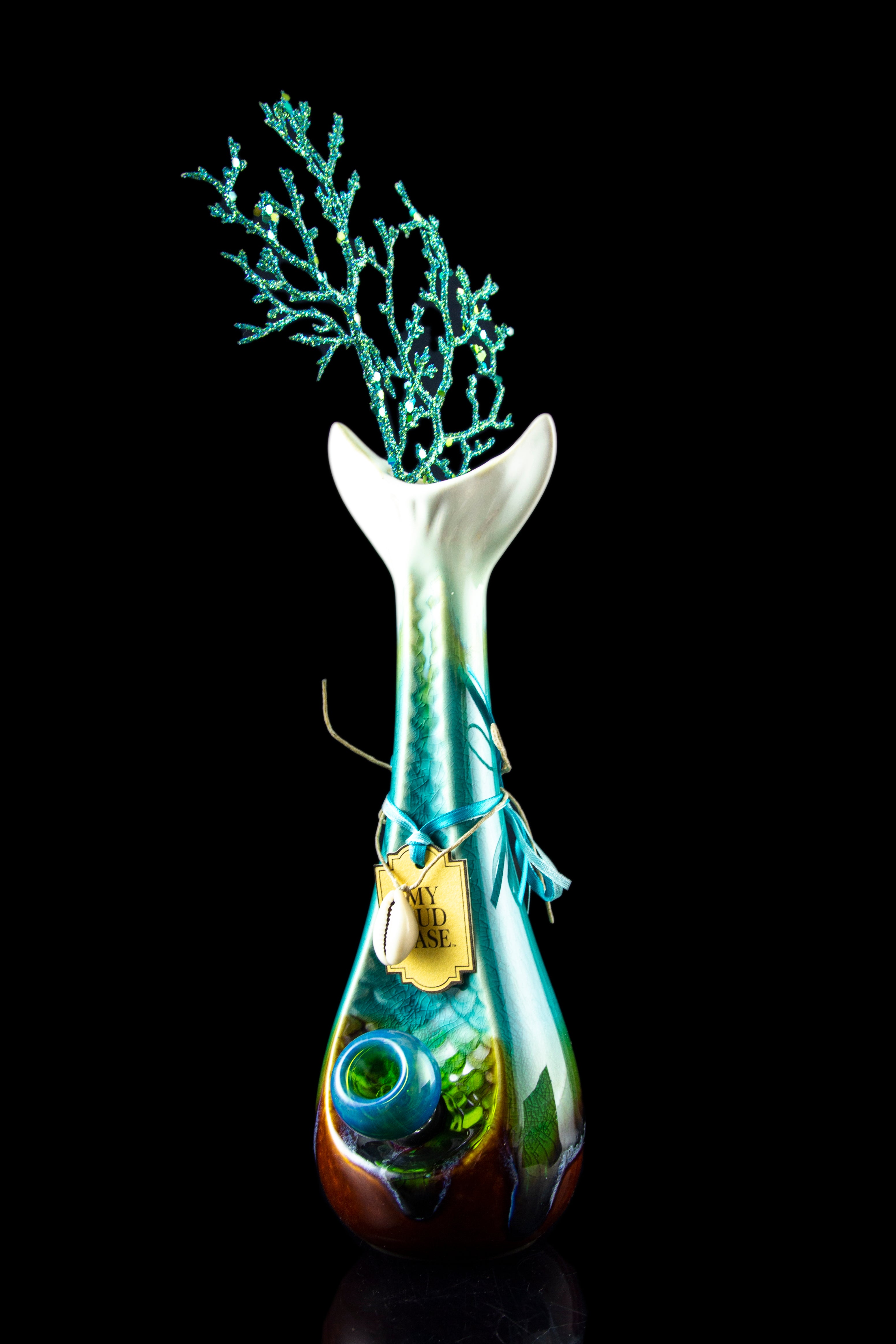 A sculpted glass water pipe with a vibrant iridescent teal and emerald finish, adorned with a miniature, glittering blue coral-like structure at the top and a decorative tag.