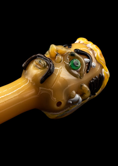Hand-blown glass cannabis pipe sculpted as a grotesque face with a single green eye and swirling amber details.