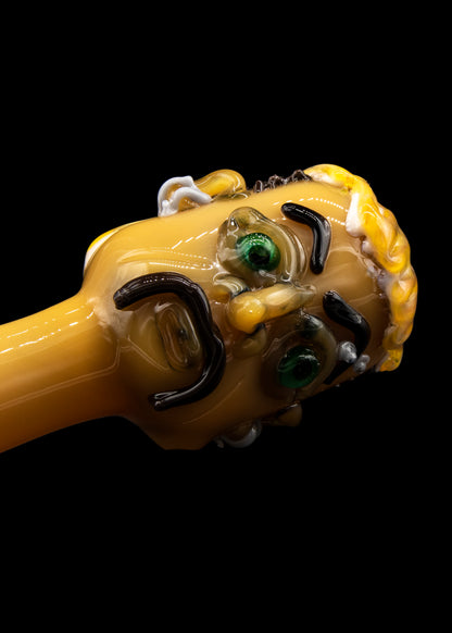Hand-blown glass pipe shaped like a cartoon character's head, featuring green eyes and a yellow and black color scheme, against a black background.