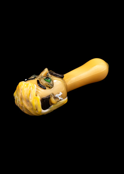 Hand-blown glass pipe sculpted as a grotesque, smiling face with green eyes, against a black background.