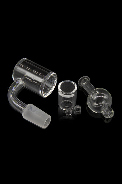 Clear glass dab rig components including a quartz banger, carb cap, and small glass bubbler, displayed on a reflective surface against a black background.