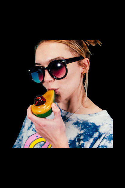 Close-up of a woman in sunglasses holding and using the Goody Avocado Hand Pipe, showing its yellow-green glass shape and red bowl.