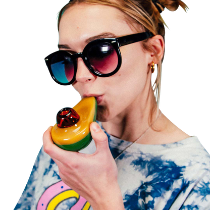 Close-up of a woman holding and bringing the Goody Avocado Hand Pipe to her lips against a teal backdrop, wearing dark sunglasses and a tie-dye shirt.