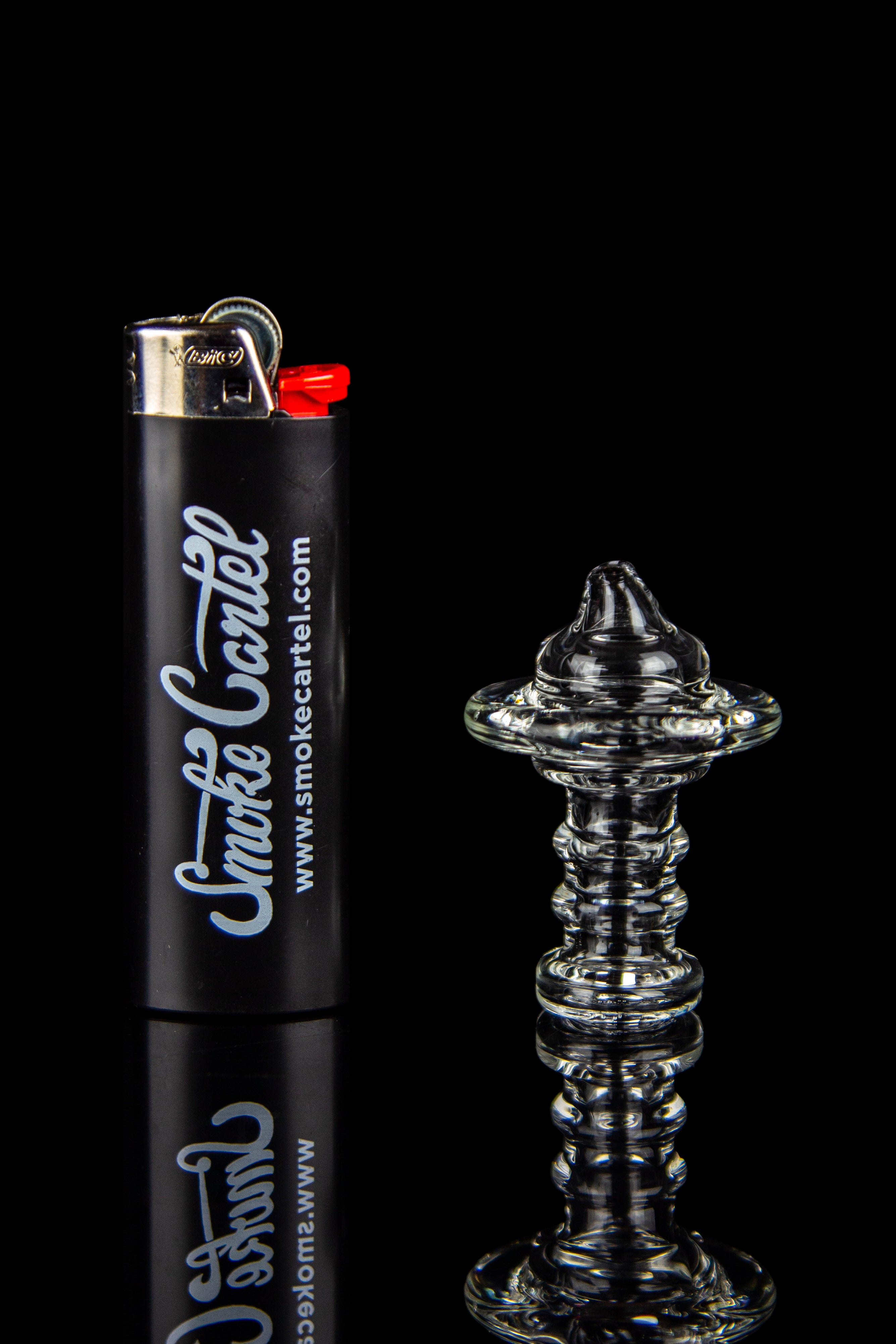 Clear glass oil rig with a black lighter branded “Smoke Cartel” reflected on a shiny surface.