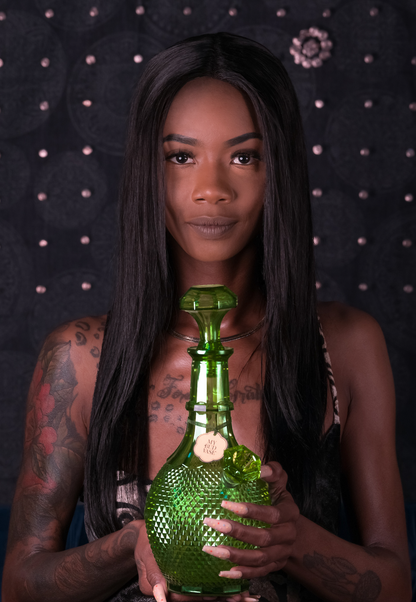 A Black person with long, straight hair and visible tattoos holds a green glass bottle with a label reading “My Bud Vase” against a dark, textured background.