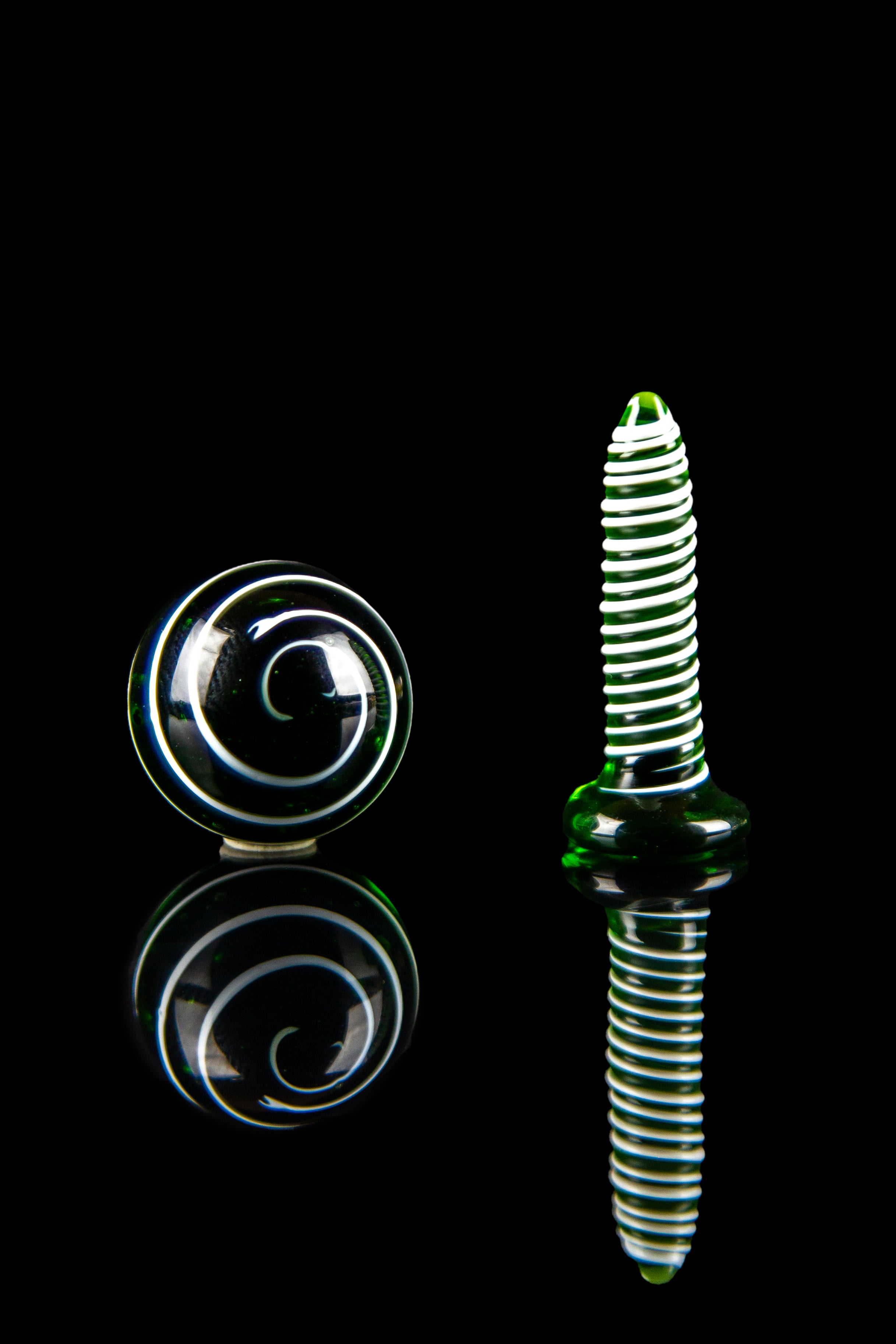 Green and clear glass cannabis oil burner and spiral marble against a black reflective surface.