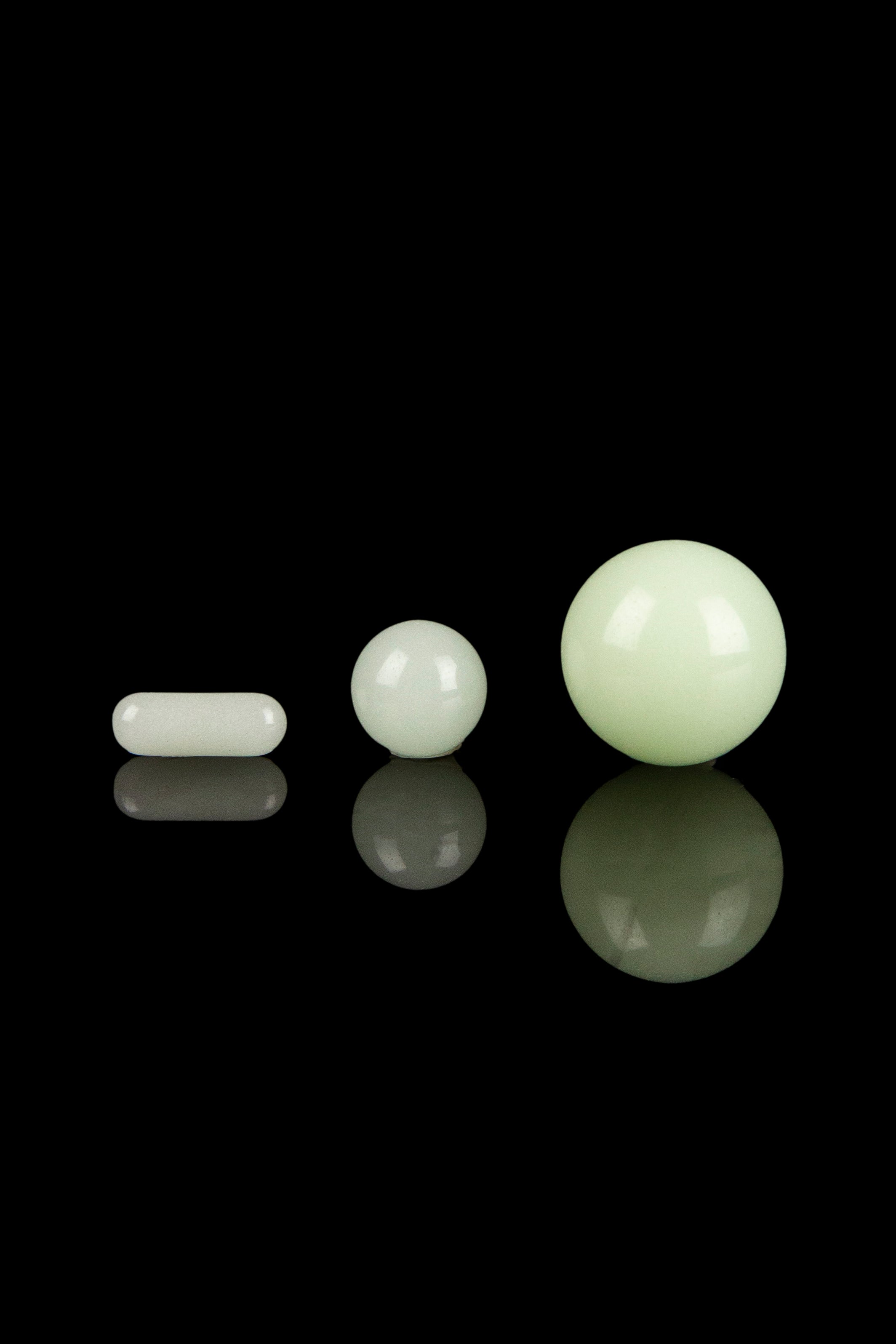 Pale green cannabis capsules and spherical concentrates displayed on a black reflective surface.