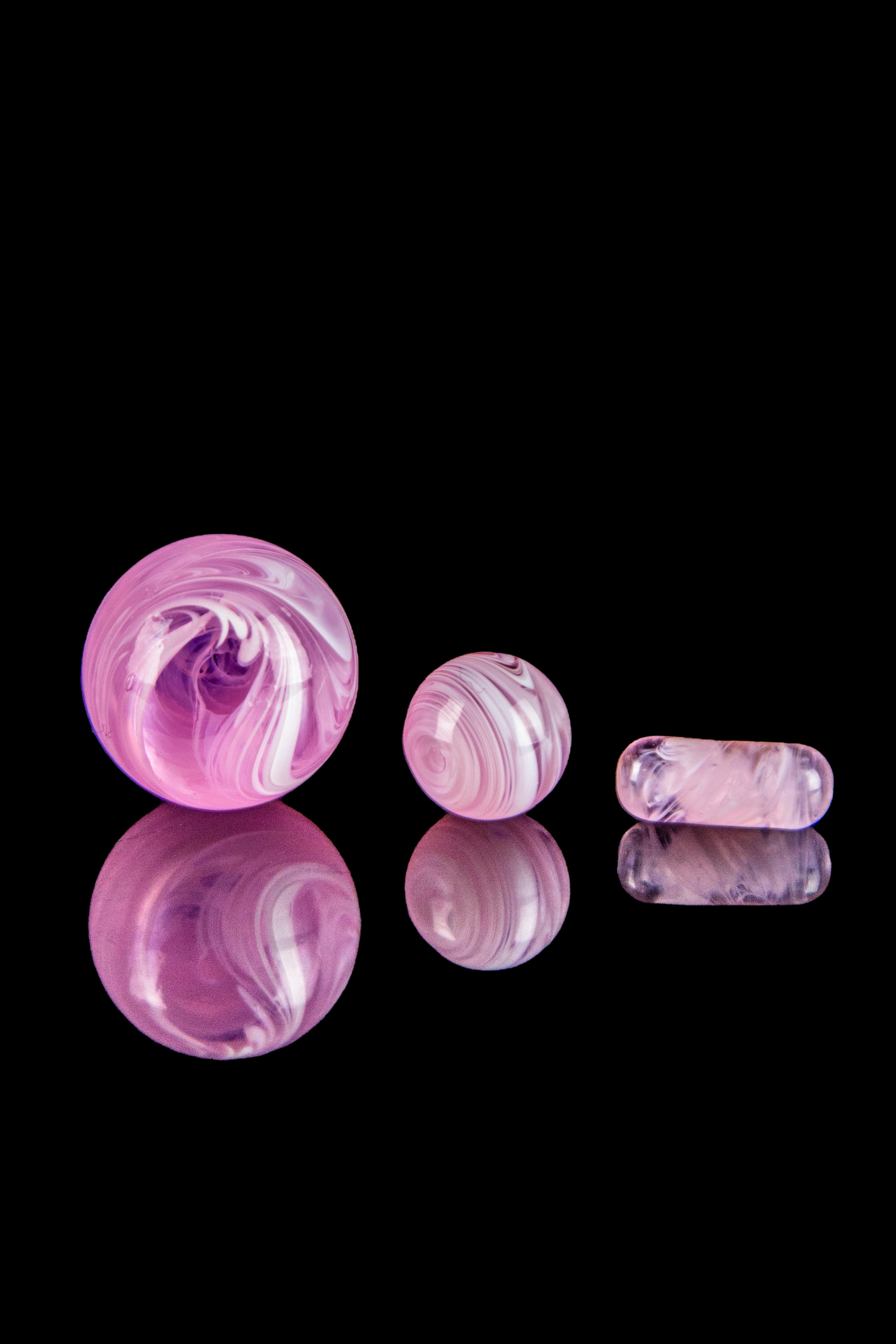 Pink and white swirled glass marbles and cylindrical pieces reflected on a black surface, likely cannabis accessories.