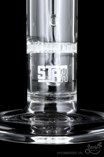 Close-up of a clear glass water pipe with “STAX” etched on the side, filled with water and reflecting on a dark surface.
