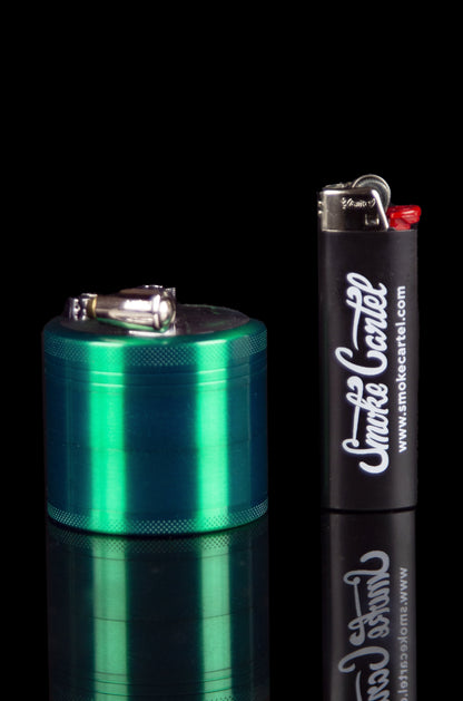 Emerald green metal grinder and lighter with “Smoke Cartel” branding, reflecting on a glossy surface.