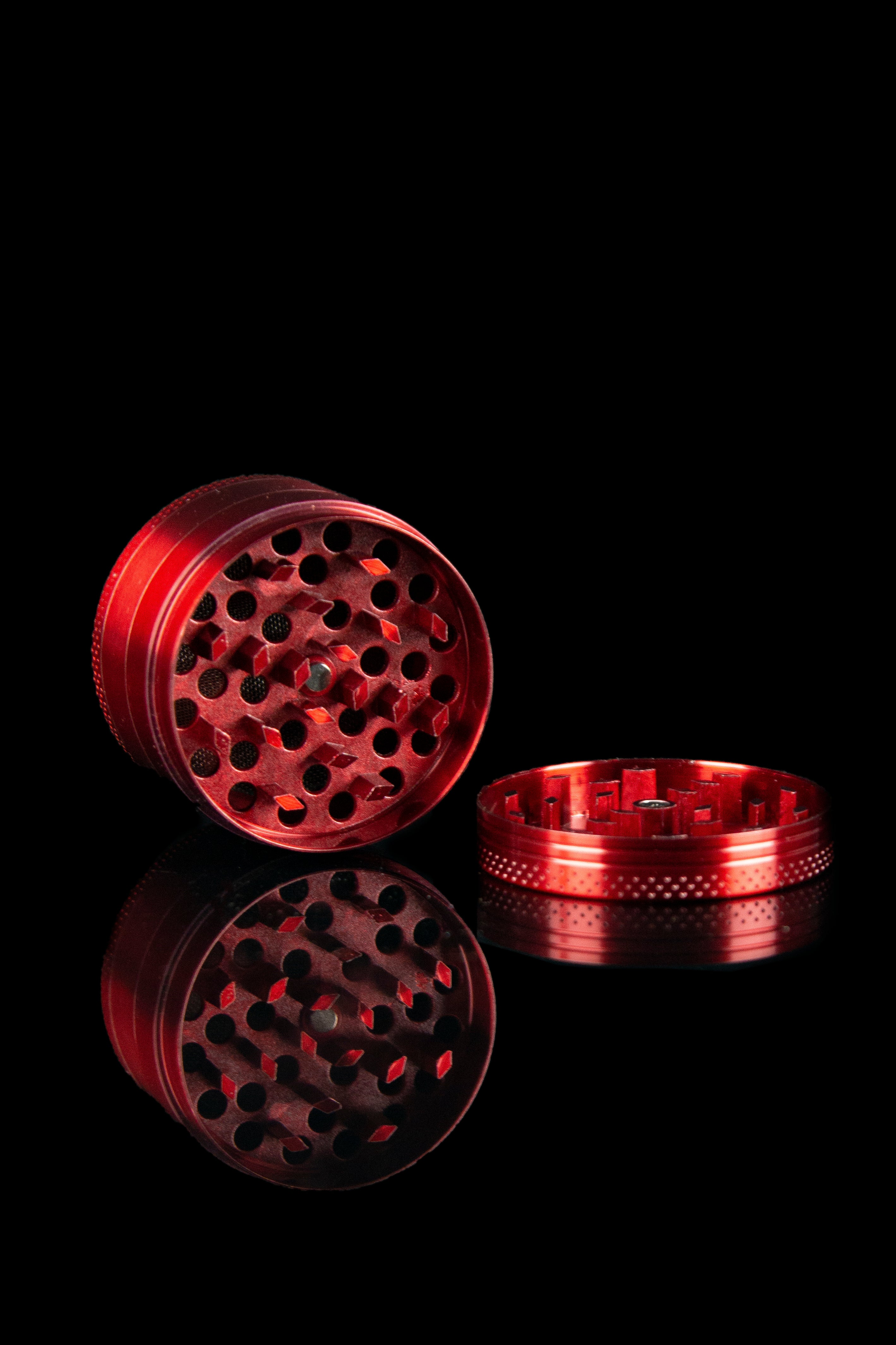 Red metal cannabis grinder with separated parts, reflecting on a dark surface.