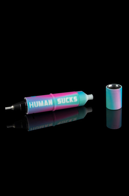 Iridescent cannabis oil syringe and accompanying mouthpiece reflected on a black surface, text reads "HUMAN SUCKS."