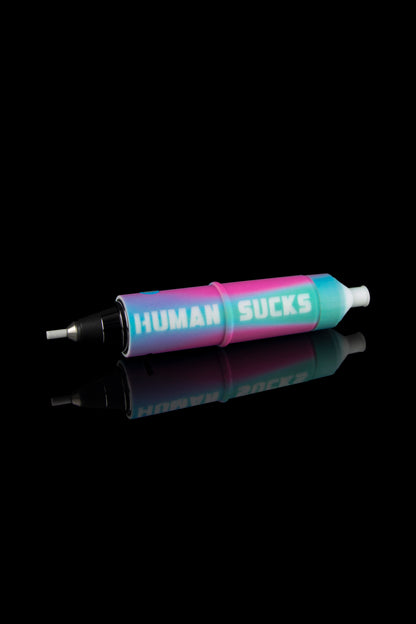 A colorful cannabis oil syringe with “HUMAN SUCKS” printed on the side, reflected on a glossy black surface.