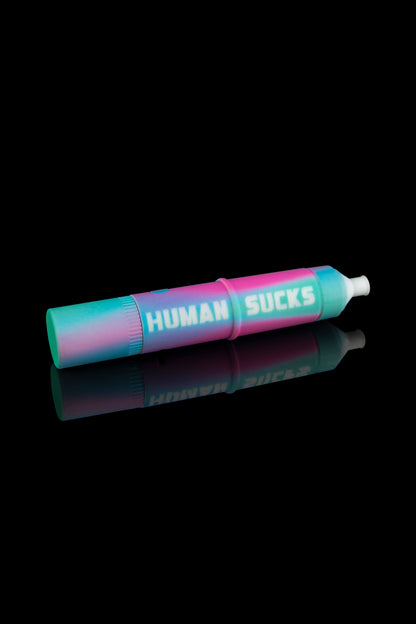 A vibrant pink and blue cannabis oil cartridge with “HUMAN SUCKS” printed on its transparent window, reflected on a black surface. 