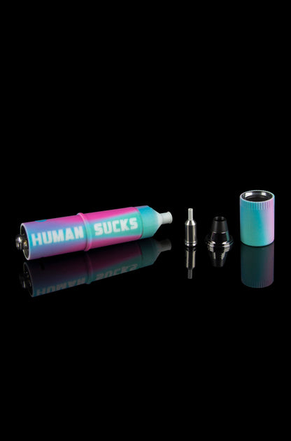 A disassembled cannabis oil vape pen with a colorful, gradient finish and the phrase “HUMAN SUCKS” printed on its side, displayed on a reflective surface against a black background.
