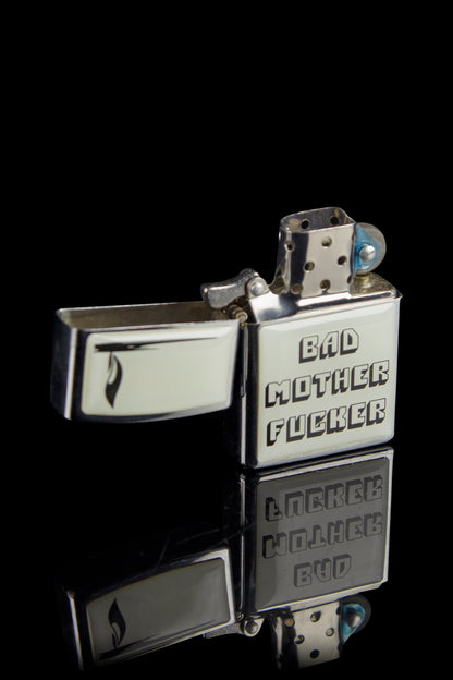 Silver Zippo-style lighter engraved with cannabis-themed profanity and imagery against a black background.