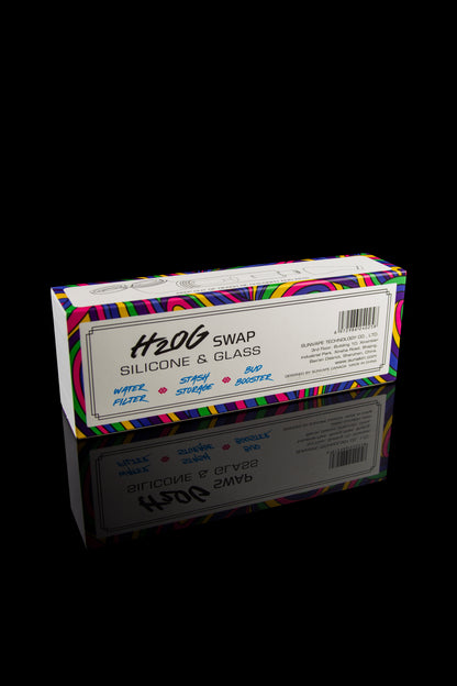 A rectangular box for cannabis accessories with colorful geometric patterns, labeled "H2OG Swap Silicone & Glass" and listing features like "water filter," "stash storage," and "bud booster," reflected on a glossy black surface.