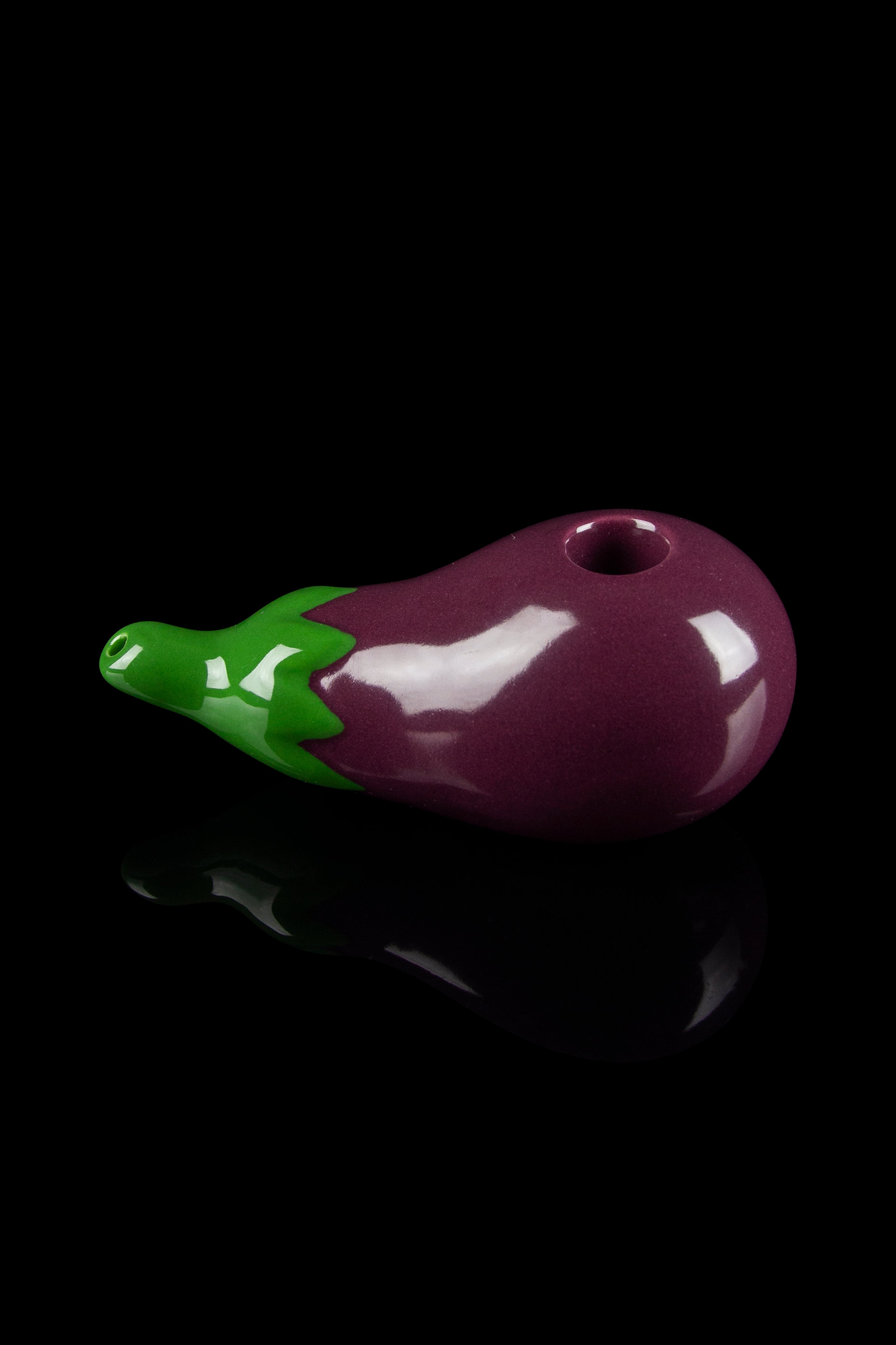 Purple glass pipe shaped like an eggplant with green leafy top, studio shot on a reflective black surface.