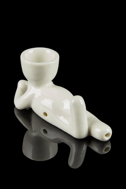 White ceramic chillum pipe shaped like a reclining figure, reflecting on a dark surface.