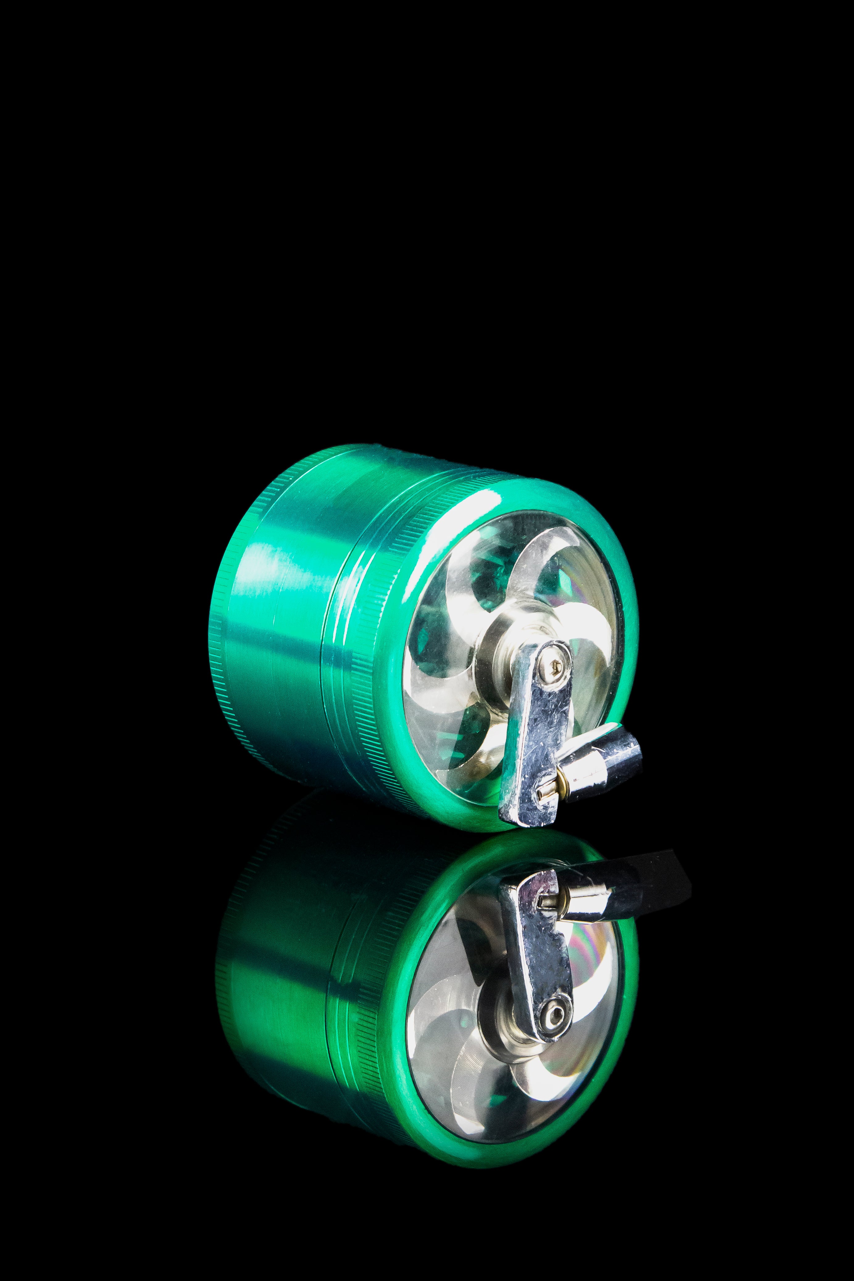 Green metal cannabis grinder with a clear acrylic chamber, displayed with its reflection on a black surface.