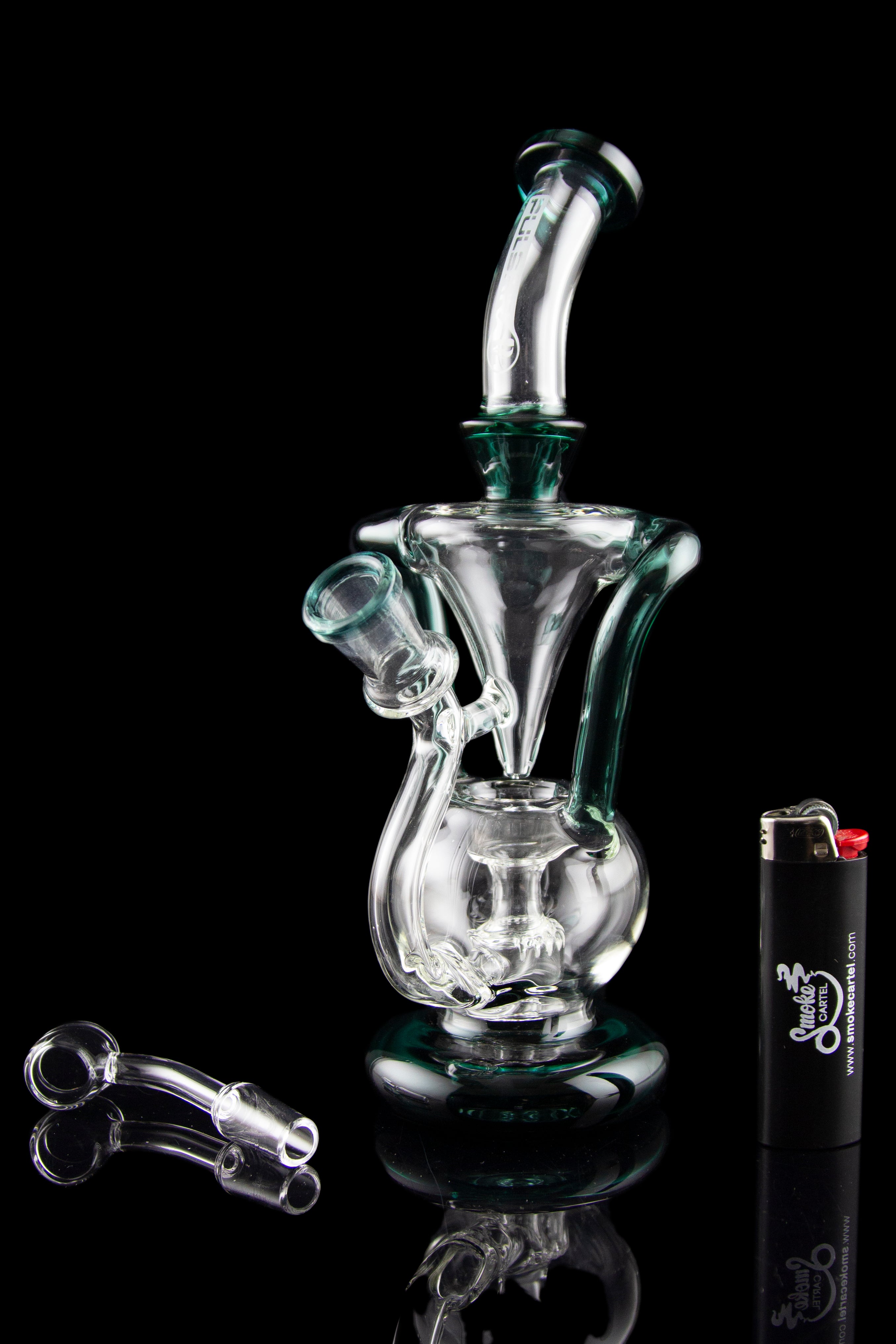 A clear glass water pipe (rig) with a unique sculptural design, alongside cannabis oil concentrate dab tools and a lighter, all reflected on a black surface.