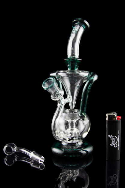 A clear glass water pipe (rig) with a unique sculptural design, alongside cannabis oil concentrate dab tools and a lighter, all reflected on a black surface.