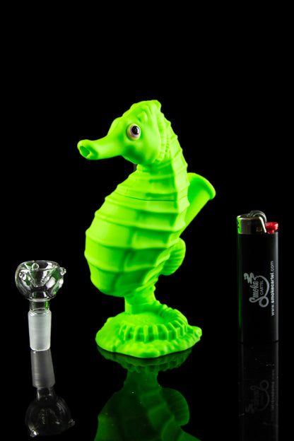 A vibrant green glass seahorse sculpture displayed alongside a glass smoking bowl and lighter, reflecting on a glossy black surface.