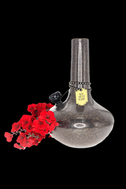 A decorative glass cannabis water pipe ("My Bud Vase") with a textured black and gold finish, displayed with red flower blossoms against a black background.