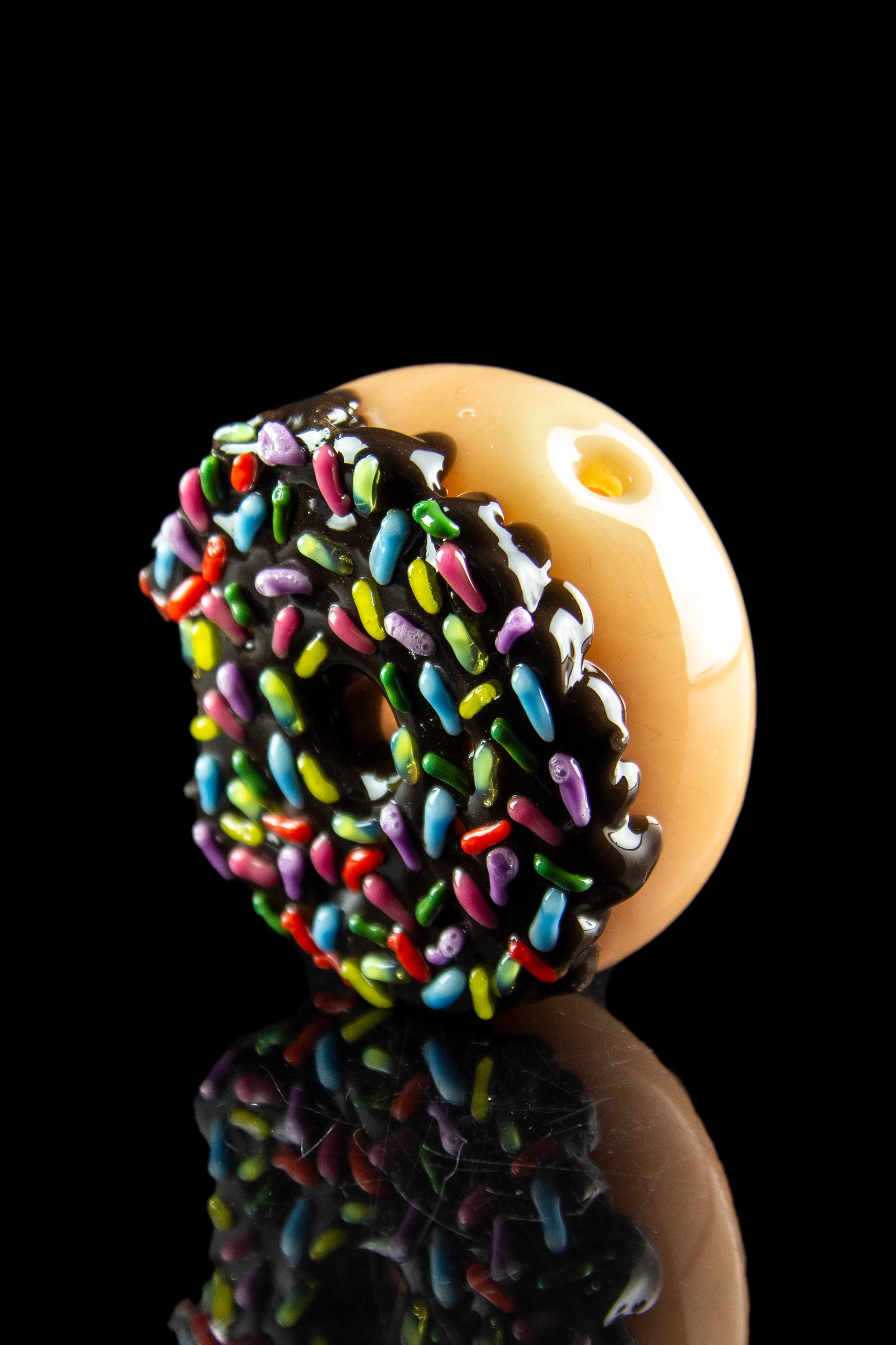 Glass cannabis pipe shaped like a frosted donut with colorful sprinkles, reflecting on a black surface.