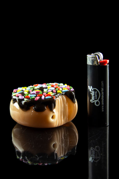 Glass pipe shaped like a donut with colorful sprinkles, alongside a lighter, reflecting on a glossy black surface.