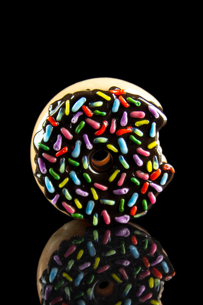 Glass pipe shaped like a chocolate donut with colorful sprinkles, reflected on a black surface.