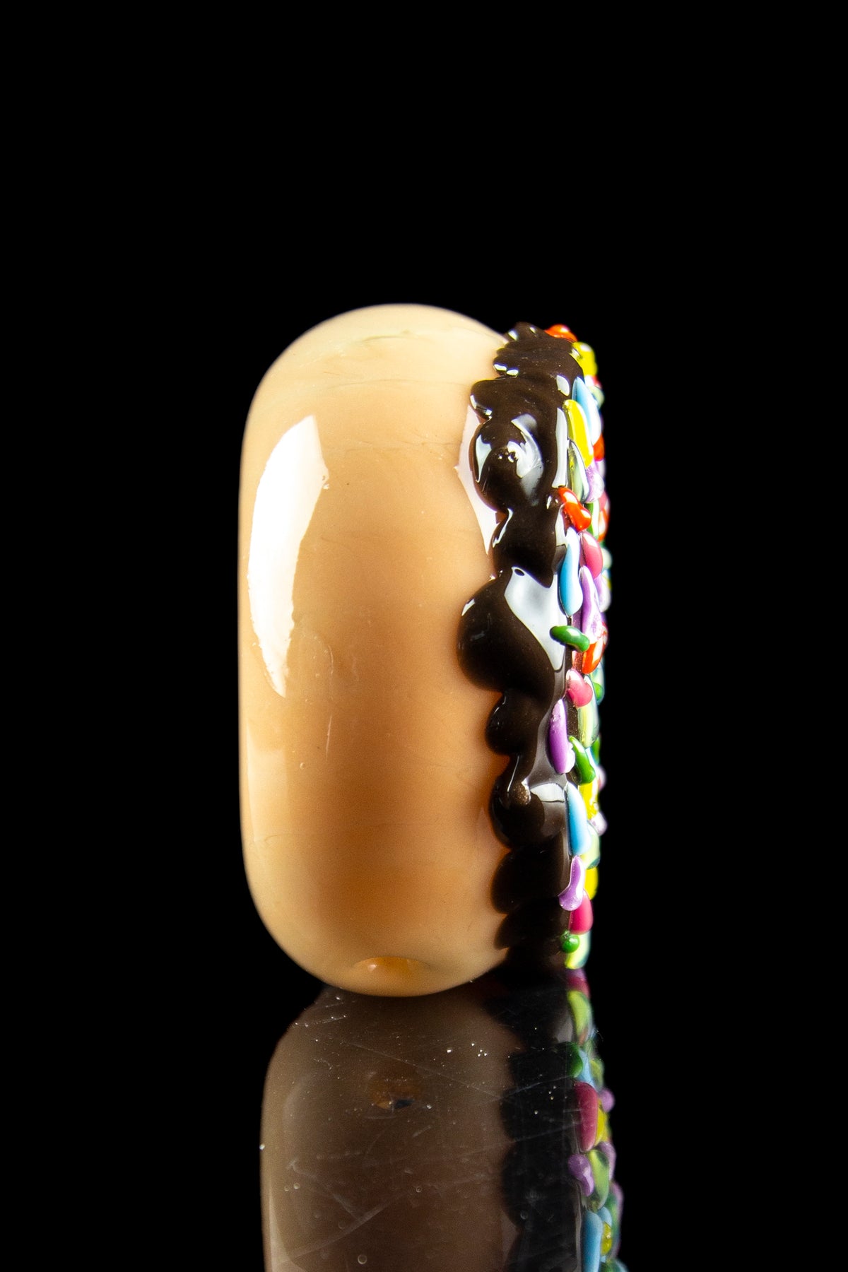Glass dab rig shaped like a frosted donut with colorful sprinkles, reflecting on a black surface.