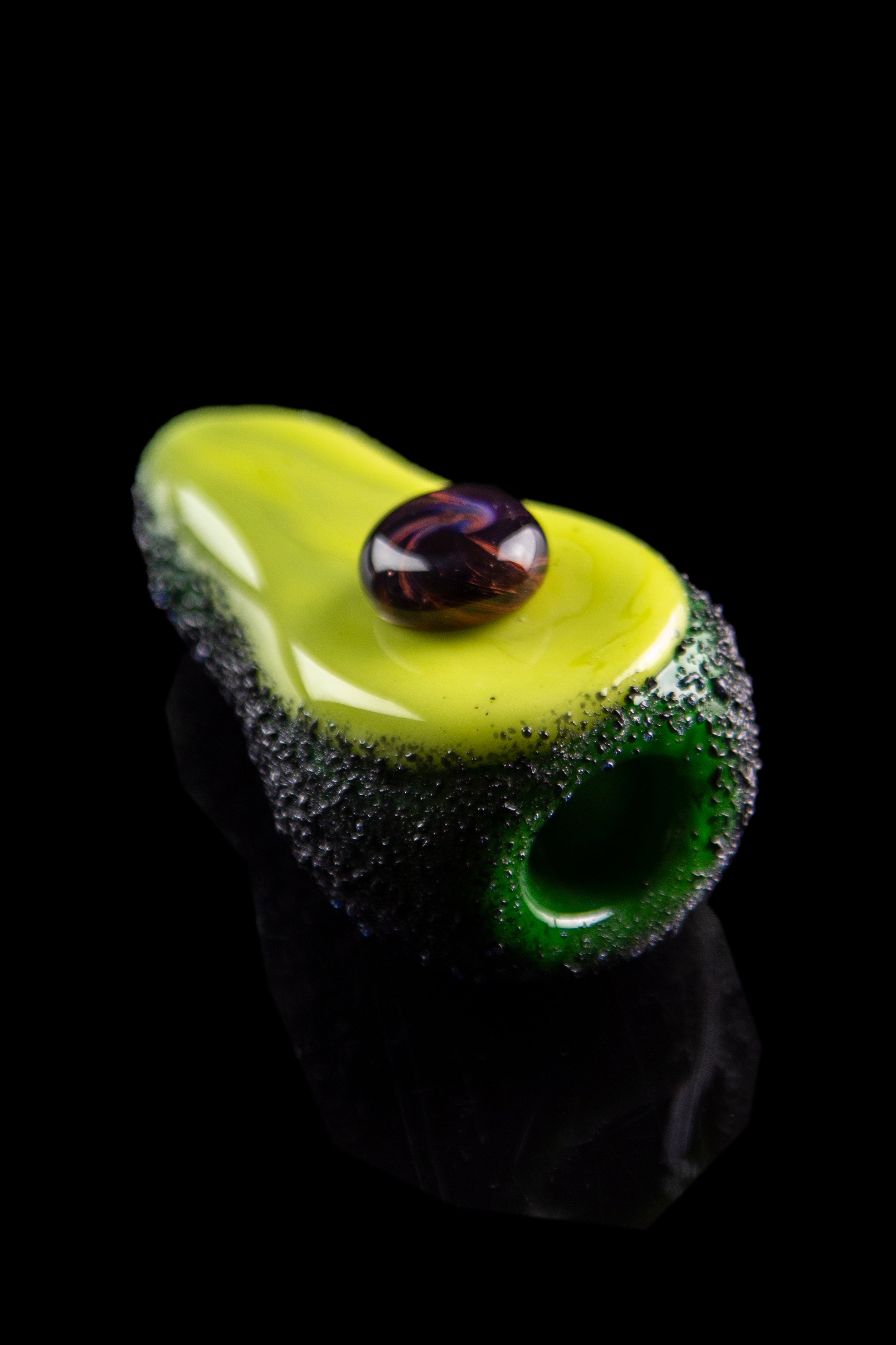 Hand-blown glass avocado-shaped pipe with a dark pit and green mouthpiece, isolated on black.