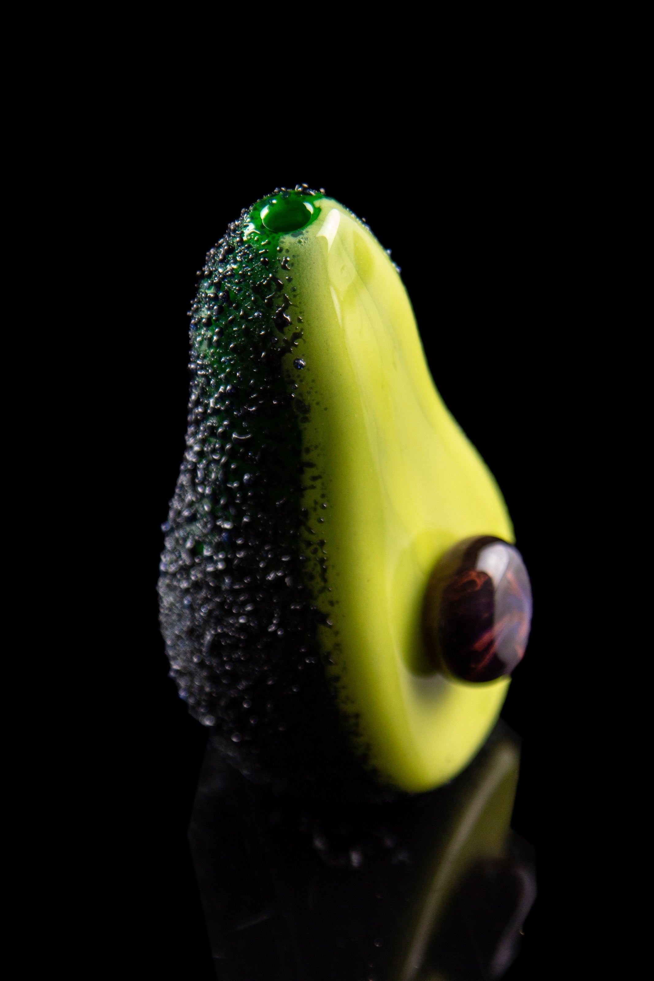 A vibrant green glass avocado-shaped piece atop a black water pipe, covered in water droplets against a dark background.