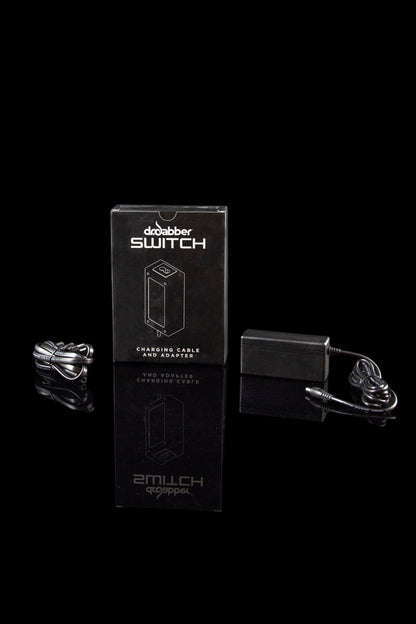 Front-facing shot of the Dr. Dabber SWITCH Replacement Charging Cable and Adapter box with coiled cable and black adapter on a reflective black surface