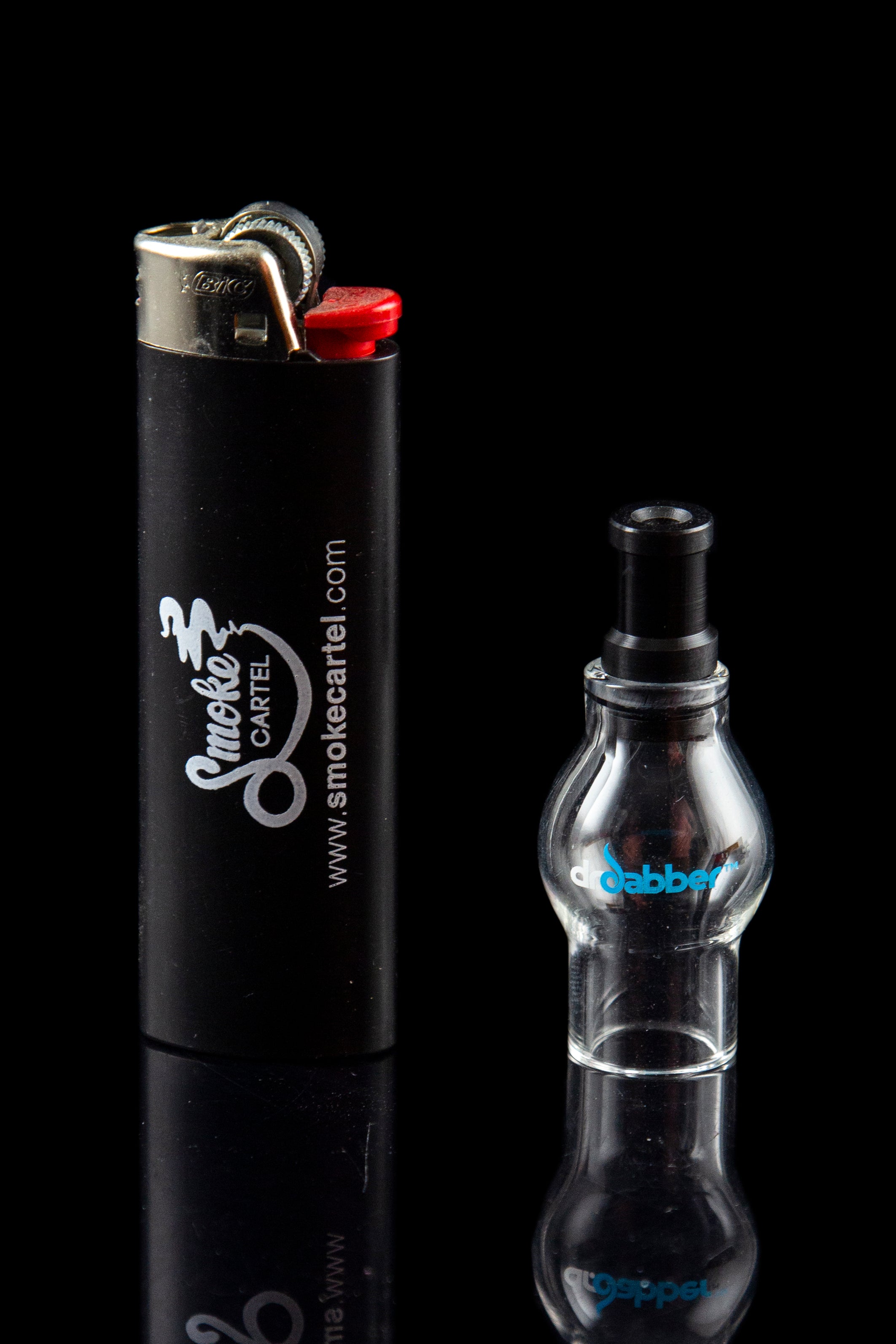 Black lighter with “Smoke Cartel” branding next to a clear glass dab rig on a reflective surface.