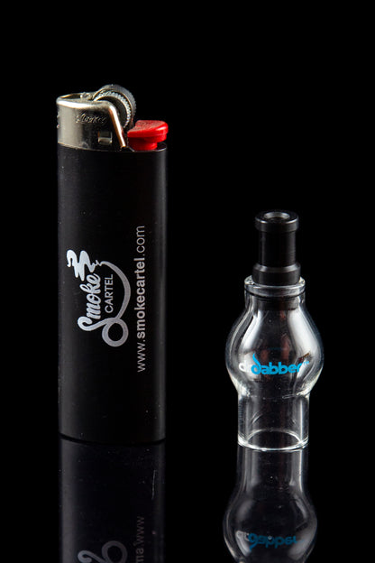 Black lighter with “Smoke Cartel” branding next to a clear glass dab rig on a reflective surface.