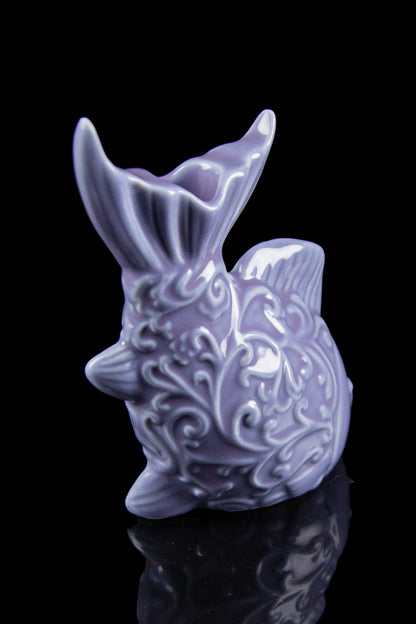 Purple ceramic pipe shaped like a pufferfish, detailed with swirling patterns, displayed on a reflective surface.
