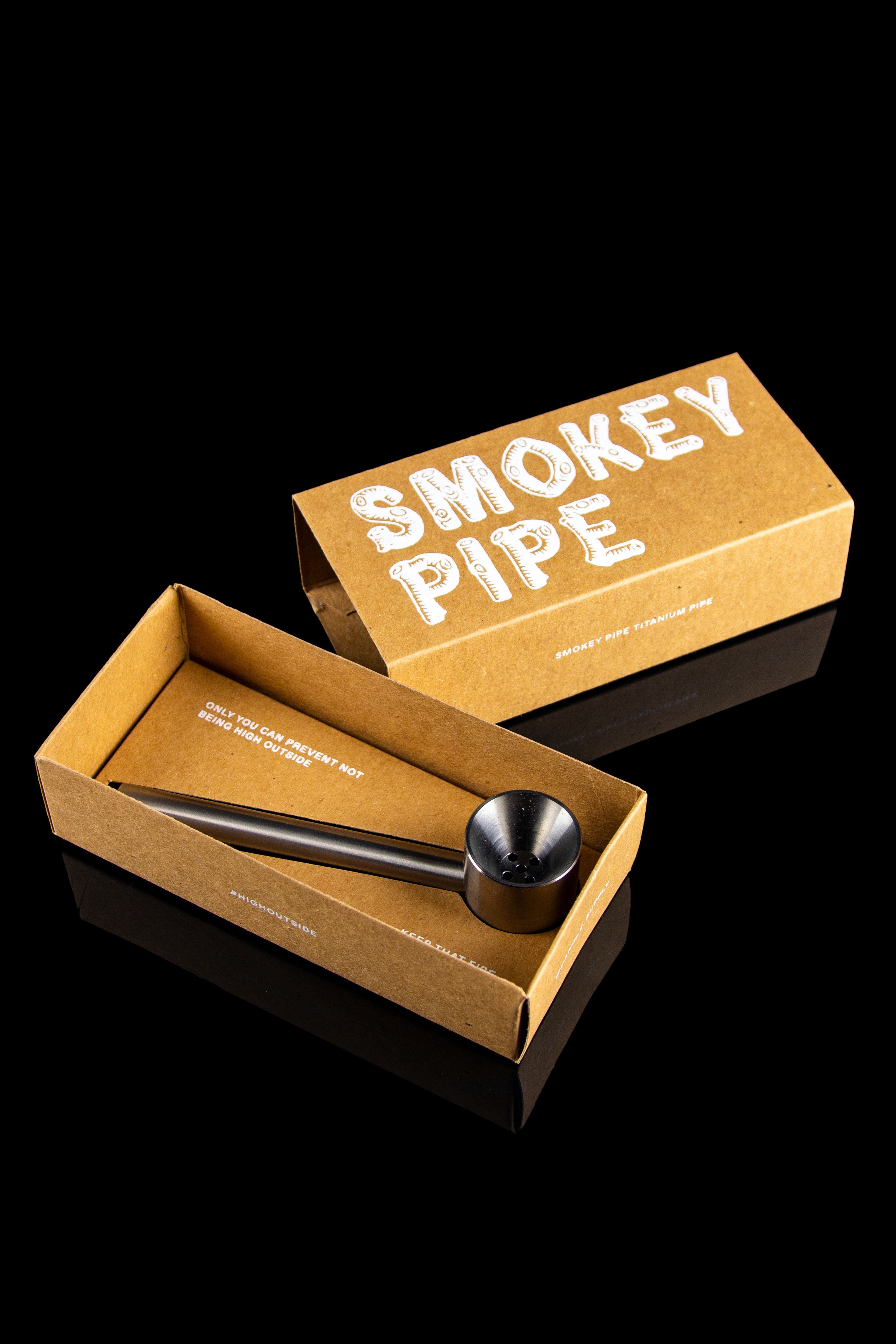 Open cardboard box displaying a black titanium smoking pipe with text reading "Smokey Pipe" and "Only you can prevent getting high outside."