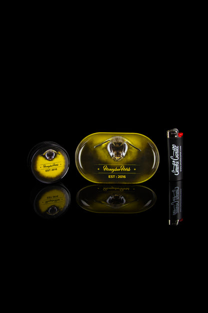 Product shot of Honeybee Herbs cannabis tins and rolling paper packaging on a reflective black surface.