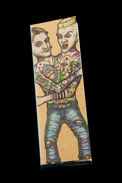 A rectangular cardboard box featuring a colorful, illustrative tattoo-style design of two men with extensive body art, likely packaging for cannabis accessories.