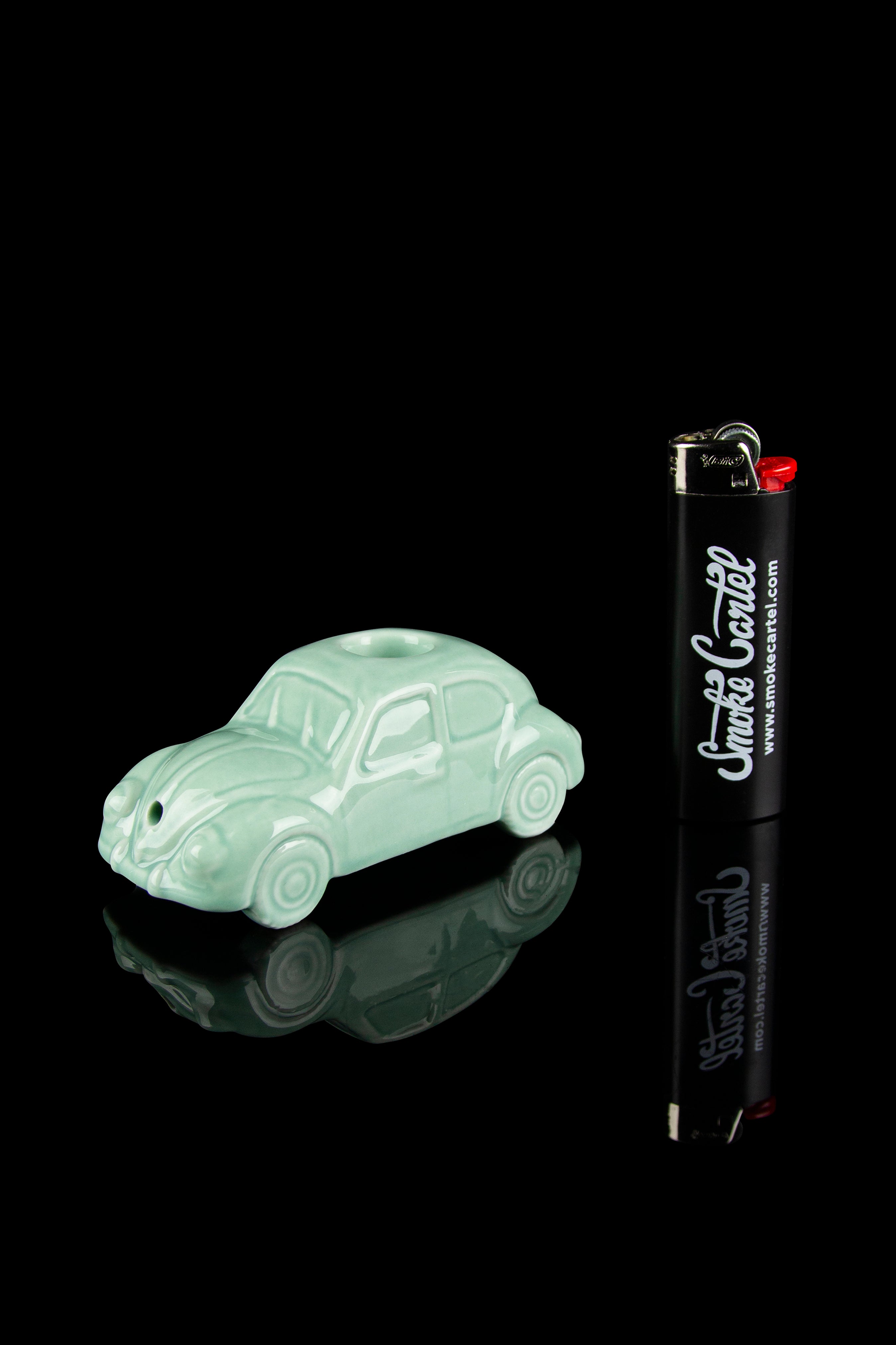Mint green ceramic Volkswagen van with a black lighter branded "Smoke Cartel" beside it, on a reflective black surface.